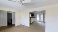 This 2-floor property offers a thoughtfully designed open-concept main level that seamlessly connects the living and dining areas to the kitchen. The living area features neutral-toned carpet flooring, crisp white walls, and a ceiling fan with a light fixture, providing both comfort and functionality. A floor vent indicates a central HVAC system throughout the space.

The kitchen is partially open to the main living area, separated by a raised breakfast bar with dark countertops and dark cabinetry, creating a natural visual contrast against the light walls. A pendant light hangs above, adding a focused point of illumination. The kitchen also features a window, allowing natural light to filter in.

Adjacent to the kitchen, a bright secondary living or dining space is highlighted by elegant French doors with large glass panes, offering views of the outdoors and filling the room with natural light. Neutral carpeting continues throughout, maintaining a consistent aesthetic across the main level. The overall ambiance is clean, neutral, and move-in ready, providing a versatile canvas for personalization across both floors.