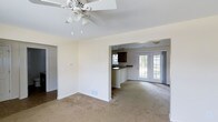 This 2-floor property offers a thoughtfully designed open-concept main level that seamlessly connects the living and dining areas to the kitchen. The living area features neutral-toned carpet flooring, crisp white walls, and a ceiling fan with a light fixture, providing both comfort and functionality. A floor vent indicates a central HVAC system throughout the space.

The kitchen is partially open to the main living area, separated by a raised breakfast bar with dark countertops and dark cabinetry, creating a natural visual contrast against the light walls. A pendant light hangs above, adding a focused point of illumination. The kitchen also features a window, allowing natural light to filter in.

Adjacent to the kitchen, a bright secondary living or dining space is highlighted by elegant French doors with large glass panes, offering views of the outdoors and filling the room with natural light. Neutral carpeting continues throughout, maintaining a consistent aesthetic across the main level. The overall ambiance is clean, neutral, and move-in ready, providing a versatile canvas for personalization across both floors.