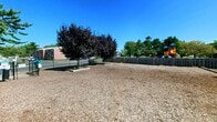 This expansive outdoor space in Bensalem offers multiple recreational areas designed for community use. A large, wood-fenced playground dominates the center, filled with wood chips for safety. Adjacent, a separate dog park provides a dedicated space for pets. The area is surrounded by mature trees, including distinctive pine and deciduous varieties, offering natural shade and greenery. Wide open spaces with well-maintained grass and dirt areas allow for various activities. Paved pathways connect different sections, ensuring accessibility. The space includes amenities such as benches and waste receptacles, catering to visitors' needs. This multi-functional outdoor area serves as a hub for community recreation and socialization.