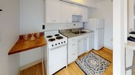 Located at 100 Memorial Dr in Cambridge, MA, this residence offers a thoughtfully laid-out living environment across its floors, connected by an interior staircase with a classic wood-railed banister. The main level features a generously sized living area with soft wall-to-wall carpeting and a clean, neutral color palette that creates an open, airy atmosphere. A pass-through window connects the living area to the kitchen, which is equipped with white cabinetry, an electric range, a full-size refrigerator, a stainless steel sink, and a breakfast bar with hardwood countertop — an efficient and functional setup. The upper level hosts well-proportioned bedrooms, each finished with plush carpeting and crisp white walls. Multiple windows throughout the rooms invite natural light and frame views of the surrounding Cambridge cityscape, including neighboring brick buildings and balconies. The bathroom is finished with tile flooring and features a shower-tub combination with full wall tiling. The overall design is clean and understated, with a bright, minimalist aesthetic that offers a versatile canvas for personal style in a prime Cambridge location.