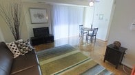 This single-floor space offers a welcoming and functional open-plan layout that seamlessly connects the living and dining areas. The living area is anchored by a sleek, dark media console supporting a flat-screen television, complemented by a plush sofa and a striped area rug in earthy tones of green, blue, and grey that adds warmth and texture to the polished hardwood flooring beneath. A framed botanical wall print adds a subtle decorative touch above the media unit. The dining area is positioned adjacent to the living space and features a compact round table paired with classic wooden chairs, ideal for intimate gatherings. A ceiling fan overhead provides both comfort and light to the shared space. Floor-to-ceiling vertical blinds line a large sliding glass door that floods the interior with generous natural light while offering a connection to the outdoors. The overall ambiance is clean, casual, and comfortable, with a neutral color palette and practical furnishings that create an inviting, livable atmosphere throughout.