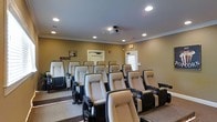 This single-level screening room is designed to deliver an authentic theater experience in a comfortable, well-appointed setting. The space is arranged with multiple rows of tiered, recliner-style seats upholstered in a neutral beige fabric and mounted on sturdy dark bases, each equipped with built-in cup holders for added convenience. The tiered layout ensures unobstructed sightlines from every seat in the room.

A ceiling-mounted projector anchors the viewing setup, positioned centrally to deliver a clear, wide projection onto the screen at the front of the room. Recessed lighting is distributed evenly across the smooth ceiling, which features classic crown molding along the perimeter, adding a polished architectural detail. The walls are finished in a warm tan tone that complements the seating and contributes to a cozy, cinematic ambiance.

Subtle LED accent lighting runs along the base of the seating rows, enhancing visibility along the aisles while reinforcing the theater atmosphere. Two windows along the rear wall provide ventilation and a connection to natural light when desired. Decorative film-themed signage on the wall adds a playful nod to the room's purpose, completing the immersive screening environment.