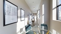 This single-floor space offers a sleek, modern aesthetic characterized by clean lines and a bright, welcoming atmosphere. Natural light streams in through tall, narrow windows lining one side of the room, casting crisp patterns across the polished concrete floors and illuminating the neutral white and gray walls throughout. The elongated layout creates a sense of depth, with distinct functional zones visible from end to end.

At the forefront, a round glass-top dining or meeting table is surrounded by teal upholstered chairs, providing an elegant and practical gathering spot. Further along, a dedicated workspace features a wooden desk paired with a white ergonomic chair and a wall-mounted adjustable task lamp, making it well-suited for focused individual work. Framed architectural drawings and floor plans adorn the walls, adding a professional and purposeful character to the space.

The elevated ceiling height enhances the open, airy feel, while flush and pendant light fixtures supplement the natural light for consistent illumination throughout the day. Overall, the space strikes a balance between functionality and contemporary style, making it versatile for both professional and collaborative use.