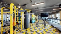 This single-floor fitness facility is designed with both functionality and visual energy in mind. The space is anchored by a striking yellow Synrgy360 functional training rig, complete with a heavy punching bag, suspension straps, and a variety of resistance attachments, making it a versatile centerpiece for full-body workouts. A collection of stability and medicine balls in assorted sizes adds further variety to the training options available.

The flooring features a bold yellow, grey, and white chevron pattern that reinforces the facility's dynamic aesthetic while providing durable, impact-absorbing surface underfoot. Benches and additional strength equipment are positioned along the perimeter, offering a well-organized layout that maximizes usable floor space.

Overhead, the industrial-style ceiling is fitted with bright linear LED lighting that ensures the entire space is well-illuminated. A large flat-screen display is mounted prominently on one wall, ideal for streaming workouts or fitness content. The open floor plan and high ceiling contribute to an airy, energetic atmosphere well-suited for a wide range of training activities.