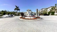 This single-story property makes a strong first impression through its well-designed entrance roundabout, which serves as both a functional traffic feature and a decorative focal point. At the center of the roundabout stands a striking large-scale metal sculpture, adding an artistic and modern touch to the arrival experience. Surrounding the sculpture is a carefully maintained circular garden bed filled with vibrant orange and purple seasonal flowers, accented by ornamental grasses and tropical red-leafed plants.

The entrance is paved with herringbone-pattern interlocking pavers, lending a polished, upscale aesthetic to the approach. Tall palm trees frame the scene, reinforcing the property's warm, Florida-inspired character. The main building, visible in the background, features a low-pitched roof, neutral stone or stucco exterior, and a clean architectural profile consistent with contemporary Florida construction. A multi-story residential building is also visible to the right, suggesting the property is part of a broader community. The overall exterior ambiance is bright, open, and well-maintained, reflecting a commitment to curb appeal and thoughtful landscaping throughout.