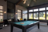 Located at 12000 W Fairview Ave in Boise, Idaho, this single-floor amenity space combines recreational and social functions within a thoughtfully designed interior. The room is anchored by soaring vaulted ceilings accented with exposed wood beams and a decorative pendant chandelier, lending architectural character and an airy sense of volume throughout.

A full-size pool table with a teal felt surface occupies the central area, positioned beneath the chandelier and complemented by a shuffleboard table along one wall. A bank of wide, multi-pane windows along the back wall floods the space with natural light and provides views of the surrounding landscaping. A flat-screen television is mounted on the opposite wall, enhancing the entertainment-focused atmosphere.

Lounge seating areas are thoughtfully distributed throughout the space. Plush navy velvet chairs with gold-toned frames are paired with circular side tables and round pedestal dining tables, offering comfortable spots for socializing. A built-in nook features open shelving with decorative accents and a cushioned bench seat. Textured wallcovering, warm sconce lighting, and neutral carpet flooring contribute to a polished, contemporary ambiance that balances relaxed comfort with modern sophistication.