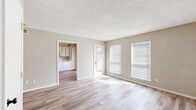 This efficiently designed single-floor unit offers a thoughtful layout across a cozy footprint, comprising a living room, kitchen, bathroom, hallway, and three closets. The living room is a bright, open space finished with light wood-look vinyl plank flooring and neutral beige walls. A textured popcorn ceiling adds character, while multiple windows fitted with horizontal blinds allow generous natural light to fill the room. A door provides direct exterior access, enhancing the sense of openness. The kitchen is positioned adjacent to the living area and features white raised-panel cabinetry extending from floor to ceiling, light stone-look countertops, a stainless steel double-basin sink, and a white dishwasher. The layout is clean and functional, offering ample storage for a unit of this scale. The bathroom is finished with a white vanity and cabinetry, a light speckled countertop, an undermount sink, and a wall-mounted mirror, creating a tidy and practical space. Consistent light wood-look flooring runs throughout the unit, unifying the spaces and contributing to the overall bright, neutral palette. Three closets provide notable storage capacity. The unit presents a clean, move-in-ready aesthetic with a simple, cohesive design throughout.