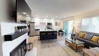 This well-appointed single-floor residence showcases a thoughtfully designed open-concept layout that seamlessly connects the kitchen, dining, and living areas. The kitchen is a standout feature, boasting white shaker cabinetry, stainless steel appliances, a bold dark-finished island with bar seating, and a contrasting black accent wall with decorative wall-mounted planters. Light wood-look flooring flows throughout the main living spaces, enhancing the bright, airy feel. The living room is anchored by a white tile fireplace with a built-in TV mount and floating shelves, while large windows allow generous natural light and views of surrounding greenery. A sliding glass door provides access to an outdoor area, further connecting interior and exterior living. The bedroom is generously sized, featuring plush carpeting, a textured accent wall, ample natural light from a large window, and room for a full dresser suite. The bathroom offers a clean, modern aesthetic with wood-look flooring, a white vanity, and a shower/tub combination. A dedicated in-unit laundry area with a stacked washer and dryer adds everyday convenience. Throughout the space, a cohesive palette of whites, blacks, and warm wood tones creates a contemporary yet welcoming atmosphere.