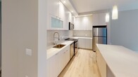 Located at 5410 McGrath Blvd in North Bethesda, Maryland, this thoughtfully designed single-floor residence offers a clean, contemporary aesthetic throughout. The open-concept kitchen is a standout feature, showcasing flat-front cabinetry in a crisp white finish, a textured tile backsplash, quartz countertops, and a central island with pendant lighting overhead. Stainless steel appliances, including a refrigerator, range, and microwave, complete the well-appointed culinary space, all set atop light-toned hardwood flooring that flows seamlessly through the living areas. The main living area benefits from expansive floor-to-ceiling windows with sleek black frames, allowing abundant natural light and offering views of the surrounding North Bethesda neighborhood. The bedrooms are comfortably sized, each featuring light hardwood floors and large windows fitted with roller shades. The primary bathroom includes a wide vanity with a undermount sink, quartz countertop, large mirror, and convenient access to a walk-in closet with built-in shelving. A second full bathroom mirrors the same refined, neutral design palette with modern fixtures and ample vanity space. Throughout the residence, neutral tones, clean lines, and quality finishes create a cohesive and inviting modern atmosphere.