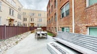 This well-appointed outdoor courtyard is situated between two distinct architectural styles — a historic multi-story red brick building with arched windows and a contemporary stucco structure — creating a visually compelling urban setting. The space is anchored by a large bluestone-tiled patio that provides ample room for outdoor dining and entertaining. A built-in outdoor kitchen station with a stainless steel gas grill set atop a stone masonry base serves as a central feature, ideal for gatherings. Adjacent to the dining area, a generously sized built-in bench with a clean, horizontal slatted design offers additional seating, complemented by a low stone fire feature with river rock detailing. Landscaping throughout the courtyard incorporates river rock beds, low shrubs, and a specimen tree, lending a natural, low-maintenance aesthetic. A paved walkway with black metal railings connects the courtyard to the street, ensuring accessible entry. Dark brown vertical privacy fencing lines the perimeter, defining the space while maintaining a sense of enclosure. The overall ambiance is that of a refined, functional outdoor retreat that blends historic character with modern amenities.