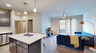 This single-floor residence is designed with a clean, contemporary aesthetic and a thoughtfully planned open-concept layout. The kitchen serves as the centerpiece of the main living area, featuring dark-toned shaker-style cabinetry, quartz countertops, a subway tile backsplash, and a full suite of stainless steel appliances including a refrigerator, range, microwave, and dishwasher. A central island with a white quartz surface provides additional prep space and seating, while pendant lighting overhead adds a polished finishing touch.

The kitchen flows seamlessly into the living area, which is bathed in natural light from a large sliding glass door that opens to a private balcony with outdoor views of surrounding greenery. A ceiling fan overhead and light wood-tone flooring throughout the main living space contribute to a bright, airy atmosphere.

The unit includes two bedrooms, each carpeted for comfort and equipped with a ceiling fan. The primary bedroom benefits from direct access to an en-suite bathroom and a walk-in closet, while the second bedroom features a window overlooking the outdoors and a dedicated closet. The overall design is cohesive, with neutral tones and warm wood accents creating a welcoming, modern environment.