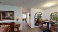 This single-floor residence offers a well-organized layout connecting multiple living spaces through a central hallway. The living area is divided into two connected zones: a front sitting room bathed in natural light from large windows dressed with decorative valances, and a more expansive main living room featuring plush carpet, framed artwork, and ample space for seating arrangements. A compact dining nook adjacent to the kitchen accommodates an intimate round table, accented by an arched decorative mirror and an ornamental metal display shelf.

The galley-style kitchen is efficiently designed with white cabinetry lining both walls, a double-basin sink, tile flooring, and standard appliances including a refrigerator and stovetop. Two carpeted bedrooms provide comfortable retreats; the primary bedroom includes a window with a decorative valance, a closet, and an ensuite bathroom featuring a single-sink vanity and a full tub-shower combination. The second bedroom is cozy and well-lit, served by an additional full bathroom with tile flooring, a single-sink vanity, and a tub-shower unit. Textured ceilings and neutral wall tones create a consistent, understated aesthetic throughout the entire space.