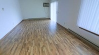 This single-floor space offers a clean and functional layout with a welcoming open feel. The main living area is finished with rich, warm-toned hardwood-style flooring that spans the entirety of the room, showcasing a varied grain pattern that adds visual depth and character to the space. The flooring is well-maintained and reflects natural light beautifully, contributing to the bright atmosphere throughout.

Natural light enters generously through a large sliding glass door dressed with vertical blinds, which also provides access to an outdoor area. The white walls keep the interior feeling fresh, airy, and neutral — an ideal canvas for personalization. A wall-mounted air conditioning unit is positioned above the sliding door, ensuring year-round climate comfort.

Baseboard heating runs along the perimeter walls, offering an efficient and consistent heat source during cooler months. The room is notably open, with no interior obstructions, maximizing usable floor space and allowing for flexible furniture arrangement. Electrical outlets are distributed at regular intervals along the walls for convenience.

Overall, the space presents a straightforward, well-lit interior with practical climate control features and durable, attractive flooring — suitable for a variety of uses and easily adaptable to different living or working configurations.