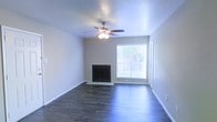 This single-story space presents a straightforward and functional design centered around a welcoming living area. The main room features dark hardwood-style flooring that runs throughout, providing a rich contrast against the neutral gray-toned walls and white baseboards. A tile-surround fireplace serves as a focal point along one wall, adding both aesthetic appeal and practical warmth to the space. Adjacent to the fireplace, a large window allows natural light to fill the room, offering a view of the greenery outside while brightening the interior. A ceiling fan with an integrated light fixture is centrally mounted, contributing to both comfort and illumination. The walls are finished with a textured ceiling treatment, adding subtle depth to the overhead surface. The overall ambiance is clean, understated, and move-in ready, with a neutral color palette that provides a versatile backdrop for a variety of furnishing styles. The open feel of the living area, combined with the natural light and classic finishes, creates a comfortable and livable environment well-suited for everyday use.