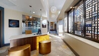 This thoughtfully designed single-floor amenity space serves as a sophisticated gathering hub with a wide range of social and recreational features. At the entrance, a refined reception desk with a warm wood and backlit gold stone facade is accented by elegant teardrop pendant lights and golden leather ottomans, setting a welcoming tone from the moment you arrive.

The main lounge area features a large sectional sofa, curated artwork, and stone tile flooring that flows seamlessly throughout the space. Soaring ceilings and expansive floor-to-ceiling windows flood the interior with natural light, while views of the surrounding urban landscape add a dynamic backdrop.

A dedicated game room includes a full-size pool table and stylish club chairs beneath generous ceiling heights and bright clerestory windows. Adjacent to this is a cozy lounge nook with built-in curved banquette seating and round café tables, perfectly positioned within a dramatic windowed bay for panoramic outdoor views.

The space also includes a coffee bar station tucked against a bold teal accent wall, and a well-appointed kitchenette featuring stainless steel appliances, dark wood cabinetry, and decorative pendant lighting. Intimate seating alcoves with patterned armchairs and game tables provide quieter spaces for conversation. Throughout, curated art pieces and cohesive finishes create a polished, contemporary ambiance.