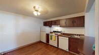 This single-floor unit in Saint Louis, Missouri, offers a straightforward, functional layout across several distinct areas. The kitchen is equipped with dark wood cabinetry providing ample upper and lower storage, a laminate countertop, a double-burner electric range, a built-in dishwasher, a refrigerator, and a single-basin sink. Wood-look flooring runs throughout the kitchen, which also features a ceiling fan with light fixture and a textured ceiling. A pass-through opening connects the kitchen area to the living space, enhancing the sense of openness between the two zones. The living area is finished with low-pile carpet and a textured ceiling, and it benefits from a sliding glass door fitted with vertical blinds that allows natural light to fill the room. A separate carpeted bedroom includes a sliding-door closet with a full-width shelf and hanging rod. The bathroom presents wood-look flooring, a vanity with a laminate countertop and mirrored medicine cabinet, a wall-mounted light bar, and a full tub-shower combination with built-in shelving. Throughout the unit, neutral wall tones and consistent finishes create a cohesive, practical living environment.