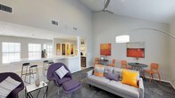 Located at 5600 Briar Oaks Ln in Raleigh, NC, this single-story space delivers a striking first impression through its open-concept layout and soaring vaulted ceilings. The generous ceiling height is accentuated by a modern ceiling fan and an arc floor lamp, contributing to a bright and airy atmosphere throughout. The main living area is thoughtfully arranged with a contemporary gray sofa accented by bold orange and yellow throw pillows, a distinctive purple lounge chair with ottoman, and a set of vibrant orange dining chairs — creating a lively, curated aesthetic. Colorful abstract artwork adorns the light gray walls, reinforcing the modern and energetic ambiance. Seamlessly connected to the living area is a kitchen and bar space, featuring a white countertop peninsula with bar seating, built-in shelving with decorative accents, and stainless steel appliances. Large windows allow natural light to filter in, enhancing the open and welcoming feel of the space. The neutral gray flooring with a patterned area rug grounds the living zone while maintaining visual continuity across the open floor plan. Overall, the space combines functional design with bold, contemporary styling in a cohesive and inviting environment.