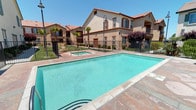 This outdoor amenity area centers around a generously sized rectangular swimming pool with clear, blue-green water, complemented by a separate spa/hot tub nearby. The pool deck is finished in large-format stone-look tiles, providing ample space for sunbathing and relaxation. Lounge chairs and a shaded dining set with an umbrella are positioned around the deck, offering both sunny and shaded seating options. A wrought iron fence encloses the pool area, ensuring privacy and security while maintaining open sightlines to the surrounding landscaping. Neatly trimmed shrubs, ornamental grasses, and tall palm trees are interspersed throughout, contributing to the resort-like atmosphere. Decorative lamp posts add architectural charm and provide evening illumination. The backdrop features multi-story residential buildings with a Mediterranean aesthetic, characterized by terracotta-tiled rooflines, warm stucco exteriors with brown trim accents, and private balconies. The overall ambiance is bright, open, and inviting, well-suited for warm-weather outdoor living under clear blue skies.