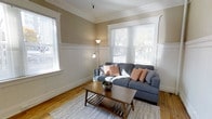 This single-floor space showcases a thoughtfully designed living room that blends classic architectural character with a warm, inviting ambiance. The room features distinctive craftsman-style wainscoting along the lower walls, painted crisp white, which contrasts elegantly with the warm taupe upper walls. Crown molding runs along the ceiling perimeter, adding a refined finishing touch that speaks to the space's traditional character.

Natural light flows generously into the room through multiple windows, including a prominent front-facing bay-style window fitted with horizontal blinds, offering views of mature trees outside. The warm-toned hardwood floors run throughout, adding richness and continuity to the space. A light gray area rug anchors the seating area, where a spacious sectional sofa in charcoal blue provides comfortable seating accented by coral-patterned throw pillows.

A mid-century modern wooden coffee table with a lower shelf serves as a functional centerpiece. A floor lamp with an adjustable arm provides additional ambient lighting. Overall, the space balances classic architectural details with a contemporary, livable aesthetic.