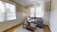 This single-floor space showcases a thoughtfully designed living room that blends classic architectural character with a warm, inviting ambiance. The room features distinctive craftsman-style wainscoting along the lower walls, painted crisp white, which contrasts elegantly with the warm taupe upper walls. Crown molding runs along the ceiling perimeter, adding a refined finishing touch that speaks to the space's traditional character.

Natural light flows generously into the room through multiple windows, including a prominent front-facing bay-style window fitted with horizontal blinds, offering views of mature trees outside. The warm-toned hardwood floors run throughout, adding richness and continuity to the space. A light gray area rug anchors the seating area, where a spacious sectional sofa in charcoal blue provides comfortable seating accented by coral-patterned throw pillows.

A mid-century modern wooden coffee table with a lower shelf serves as a functional centerpiece. A floor lamp with an adjustable arm provides additional ambient lighting. Overall, the space balances classic architectural details with a contemporary, livable aesthetic.