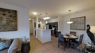 This single-floor space showcases an open-concept layout that integrates the kitchen and dining area into one cohesive living environment. The kitchen is anchored by a white peninsula with pendant lighting above, offering both additional prep space and a natural divider within the open floor plan. Dark espresso cabinetry lines the kitchen walls, providing a striking contrast against the white countertops and light-toned walls. Stainless steel appliances, including a refrigerator, microwave, and range, contribute a modern, functional aesthetic. Light wood-toned flooring extends throughout the space, creating a warm and consistent visual flow from room to room. The dining area is comfortably sized, accommodating a dark wood table and chairs beneath a statement pendant light fixture. A decorative wall accent adds a touch of character to the dining space. Recessed and flush-mount ceiling lighting ensures the interior remains bright and well-illuminated. The open sightlines throughout the space enhance the sense of spaciousness, while neutral wall tones provide a versatile backdrop. A hallway visible in the background suggests additional rooms beyond the main living area.