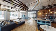 Located at 8698 E 116th St in Fishers, Indiana, this single-floor space showcases a striking modern industrial aesthetic that blends raw architectural elements with refined, contemporary furnishings. Soaring ceilings with exposed concrete, steel beams, and polished silver ductwork define the overhead landscape, while suspended greenery and decorative pendant lighting add warmth and visual interest throughout.

The open main area centers around a generously sized lounge anchored by a deep navy sectional sofa atop a textured area rug, complemented by round accent tables and curated seating arrangements. Large floor-to-ceiling windows line the perimeter, flooding the space with natural light and offering views of the surrounding streetscape.

A bar-style counter with dark tile detailing and bar seating sits adjacent to a dramatic exposed brick wall hung with Edison-style pendants, creating a distinct social zone. Glass-walled enclosures provide semi-private meeting or lounge areas without sacrificing visual connectivity to the main floor.

A reception-style corridor features a detailed dark tile desk, copper-toned pendant fixtures, and leather seating. Curated artwork displayed on slate-toned tile walls adds a gallery-like quality. The hardwood flooring ties all zones together cohesively, resulting in a sophisticated, multifunctional environment.