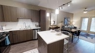 This 1-bedroom, 1-bathroom residence is thoughtfully designed with a modern aesthetic and high-quality finishes. The open-concept layout seamlessly connects the kitchen, dining, and living areas, creating a cohesive and functional living space. The kitchen is a standout feature, equipped with dark wood-toned cabinetry, a white quartz countertop island with bar seating, a subway tile backsplash, and stainless steel appliances including a dishwasher. Pendant lighting with cylindrical shades hangs above the island, adding a contemporary touch, while a track lighting system provides additional illumination overhead. The wood-look flooring runs throughout, lending warmth and continuity to the space. The dining area sits adjacent to the kitchen, offering an intimate setting for meals, and flows naturally into the living area, which is accented with warm neutral tones on the walls. The overall ambiance is clean, modern, and inviting, with a careful balance of functionality and style that makes this residence well-suited for comfortable everyday living.