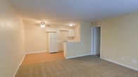 This single-floor unit presents an open-concept design that seamlessly connects the living area to the kitchen. The living space is finished with neutral-toned carpet, creating a comfortable and understated foundation for the room. A ceiling fan with integrated lighting is centrally positioned, providing both air circulation and ambient illumination.

The kitchen is set slightly apart by a half-wall partition, defining the space while maintaining visual openness to the main living area. It features white cabinetry both above and below the counter, a white refrigerator, and a sink positioned beneath upper cabinets. The kitchen floor transitions to hard-surface flooring, offering a practical contrast to the carpeted living space. White cabinetry and appliances give the kitchen a clean, cohesive appearance.

A closet with a paneled door is visible adjacent to the kitchen area, providing convenient storage. The walls throughout are painted in a soft, warm beige, contributing to the unit's bright and neutral overall ambiance. The layout is efficient and functional, making good use of the available space across the single floor.
