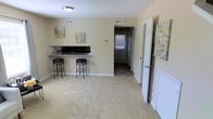 This two-floor residence offers a straightforward, practical layout designed for comfortable everyday living. The main living area is finished with wall-to-wall carpet in a warm neutral tone, creating a soft and inviting atmosphere. A built-in breakfast bar with dark granite-style countertop and two bar stools serves as a natural dividing point between the living space and the kitchen beyond, adding a casual dining option without sacrificing openness. The kitchen is partially visible through the bar opening, featuring dark appliances and ample counter space. Walls throughout are painted in a soft, warm beige, complemented by white trim and doors that keep the interior feeling bright and clean. Ceiling-mounted light fixtures provide consistent overhead illumination, while multiple HVAC vents suggest efficient climate control throughout the home. A hallway visible from the living area leads to additional rooms, with a window at its end drawing in natural light. White interior doors offer access to adjoining spaces, and a textured ceiling adds subtle visual depth. The overall ambiance is clean, neutral, and move-in ready.