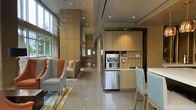 This single-floor space presents a well-appointed communal area combining lounge and kitchenette functionality within a contemporary design framework. Large floor-to-ceiling windows along one wall flood the space with natural light, highlighting the neutral-toned large-format tile flooring throughout. The lounge area is furnished with modern upholstered armchairs and sofas accented with orange and warm-toned throw pillows, anchored by a multicolored geometric area rug that adds a vibrant focal point. An elegant chandelier with a branching, crystalline design adds a refined decorative touch overhead. Recessed lighting supplements the natural light, ensuring the space remains bright and inviting. Toward the rear, a built-in kitchenette features light wood cabinetry extending floor to ceiling, paired with stainless steel appliances including a refrigerator and microwave. A counter-height seating area with light blue chairs provides an additional casual dining or gathering spot. The open corridor layout creates a sense of depth and flow, with glimpses of additional seating areas further along, making this an ideal space for socializing or relaxing.