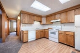 This single-floor unit offers a practical and efficiently organized layout, accommodating three bedrooms, one full bathroom, a living room, and a kitchen. Throughout the space, warm wood-paneled walls and textured drop ceilings lend a consistent, retro character, while plush carpeting runs through the living and bedroom areas.

The living room is a comfortable open space adjacent to the kitchen, featuring vertical blind-covered windows that let in natural light and a baseboard heating system along the perimeter. The kitchen is equipped with warm oak cabinetry, white laminate countertops, a double-basin stainless steel sink, and white appliances including a range, dishwasher, and refrigerator. Light wood-style flooring and a skylight panel brighten the kitchen area.

Each of the three bedrooms features carpeted floors, baseboard heaters, and windows with vertical blinds offering views of surrounding trees and the outdoors. The primary bedroom is notably spacious for the overall footprint of the unit, with wood-paneled walls and ample wall space. The additional bedrooms are cozier in scale but similarly appointed.

The bathroom features a tiled floor in a small mosaic pattern, a combined tub and shower, a toilet, and a wood-trimmed vanity with a single basin sink and mirrored medicine cabinet above.