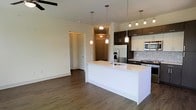 This single-floor space showcases a contemporary open-concept layout that seamlessly connects the kitchen and living areas, creating a bright and functional living environment. The kitchen is a standout feature, equipped with dark espresso cabinetry contrasted by crisp white lower cabinets and a clean white countertop island with an integrated sink. A mosaic tile backsplash adds texture and visual interest, complementing the stainless steel appliances including a refrigerator, range, and microwave. Stylish pendant lights hang above the island, enhancing both ambiance and task lighting, while a multi-bulb track light illuminates the kitchen workspace. The open living area extends from the kitchen, featuring warm wood-look plank flooring that runs throughout the space, adding a sense of continuity and warmth. Neutral wall tones keep the space feeling airy and versatile. A ceiling fan is visible in the living area, contributing to year-round comfort. The entryway features a solid wood-paneled door and is well-lit with a flush-mount fixture, offering a welcoming arrival experience. Additional doorways suggest access to private rooms, rounding out this well-appointed, modern single-level layout.
