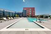 Located at 1 Green Island Blvd in Worcester, Massachusetts, this outdoor amenity space is a thoughtfully designed community environment that blends modern architecture with resort-style recreation. The centerpiece is a large, rectangular swimming pool with crystal-clear water, gradual entry steps, and an accessibility lift, surrounded by an expansive pool deck lined with chaise lounges and shaded pergola seating. The pool area is enclosed by sleek black iron fencing and offers open views of the surrounding skyline under ample natural light.

Adjacent to the pool, a welcoming courtyard features large-format tile pavers, high-top communal tables, lounge seating with lush greenery, and overhead string lights that lend an inviting ambiance. Curated garden beds with flowering plants and young trees are interspersed throughout, adding color and texture to the environment.

A manicured lawn area provides open green space framed by the modern multi-story buildings, which feature a striking combination of gray panel siding and bold red and charcoal facade elements with large windows and private balconies. Landscaped pathways connect the various zones of the space, offering a cohesive and accessible flow throughout. The overall design reflects a contemporary urban aesthetic with a strong emphasis on outdoor community living.