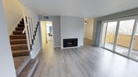 This well-appointed two-story space showcases a clean, contemporary design with a neutral color palette of soft grays and whites throughout. The main living area is anchored by a sleek black fireplace set against a crisp white surround, creating a focal point that adds both warmth and style to the room. Light wood-look flooring extends seamlessly across the open-plan layout, lending a sense of continuity and spaciousness to the interior.

Sliding glass doors along the far wall bathe the living space in abundant natural light while providing direct access to an outdoor area, enhancing the connection between indoor and outdoor living. A ceiling fan is visible in the adjoining area, adding to the practical comfort of the space.

The staircase, finished with gray carpet and clean white railings, leads to the upper floor and adds a sense of vertical dimension to the home. Neutral gray walls and white ceilings keep the atmosphere bright and airy. A hallway visible from the main living area offers a glimpse into additional rooms, suggesting a well-considered and functional floor plan. Overall, the space presents a fresh, move-in-ready aesthetic with modern finishes and a cohesive design sensibility throughout both levels.