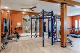 Located at 837 NW Donovan Rd in Lee's Summit, MO, this single-floor fitness facility is thoughtfully designed to accommodate a wide range of training disciplines. The main workout area is characterized by warm terracotta-orange walls, light hardwood-style flooring, and exposed wooden beam columns and ceiling trusses that lend the space an industrial yet inviting aesthetic. Ceiling fans and recessed lighting ensure a comfortable environment throughout.

The facility's standout feature is a built-in rock climbing wall, complete with colorful holds and safety ropes, paired with a cushioned crash mat — an uncommon amenity that sets this gym apart. At the center of the space, a freestanding functional training rig supports suspension trainers, cables, and other attachments for versatile strength and conditioning work. A dedicated floor mat area provides ample room for stretching, bodyweight exercises, and functional movement.

The cardio zone is well-equipped with treadmills, ellipticals, a stationary bike, and a rowing machine, all positioned near windows that allow natural light to fill the space. A separate strength area features cable machines, benches, free weights, and additional resistance equipment. Mirrors are strategically placed throughout to maximize visibility and the sense of openness.

A distinct barre studio annex, accessible via glass-panel doors, offers a clean, neutral-toned space with full-wall mirrors, mounted barre rails, and light flooring — ideal for dance fitness, yoga, or low-impact training sessions.
