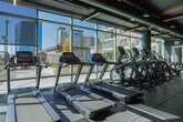Located at 902 W Peachtree St NW in Midtown Atlanta, this contemporary fitness center is designed to deliver a premium workout experience. The space features a striking industrial aesthetic, with a dark color palette, exposed ductwork, and sleek linear lighting that creates an energizing atmosphere throughout.

Floor-to-ceiling windows line one wall of the facility, flooding the space with natural light and offering expansive views of the Atlanta skyline — an inspiring backdrop for cardio sessions. A row of treadmills, ellipticals, and other cardio machines is positioned directly in front of these windows to take full advantage of the city views.

The strength training area is equally well-appointed, featuring a multi-station cable machine, adjustable benches, a full dumbbell rack, and an assortment of kettlebells, medicine balls, and exercise balls organized neatly on dedicated storage racks. Large mirrors amplify the sense of space while allowing users to monitor their form. Multiple flat-screen displays are mounted throughout the room for entertainment and on-demand fitness programming access.

Durable rubber flooring in a checkered pattern covers the entire workout area, providing both safety and functionality. A dedicated rowing machine zone offers additional variety, and a Fitness on Demand system is available for guided virtual workouts. Ceiling fans and ventilation ensure comfort during intense training sessions.