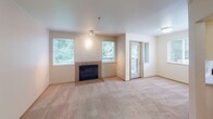 Located in University Place, Washington, this single-floor residence offers a well-organized layout with a neutral palette and functional design throughout. The living room serves as the centerpiece of the home, featuring a fireplace framed by two windows that frame lush, green wooded views. Plush carpeting extends across the space, lending warmth underfoot, while a door provides direct access to a private balcony, extending the living area outdoors.

The kitchen is efficiently designed with white cabinetry accented by warm wood trim, light countertops, and hardwood flooring. A window above the sink brings in natural light, and appliances including a dishwasher, refrigerator, and range are included. Adjacent to the kitchen, a transitional area with continued hardwood flooring connects the main living spaces to the private quarters.

The bedroom is generously proportioned, with soft carpeting, a large window overlooking greenery, and a baseboard heating unit for year-round comfort. The bathroom features hardwood flooring, a wide vanity with ample cabinetry, a large mirror with overhead lighting, and open shelving for additional storage. Throughout, the space maintains a clean, understated aesthetic suited to comfortable everyday living.