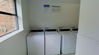This shared laundry facility is a practical, single-floor space designed to meet the everyday needs of its users. The room is clean and straightforward in its layout, featuring a row of top-loading washing machines and dryers neatly arranged along the wall, providing ample capacity for multiple loads at once. The walls are finished in a neutral tone, creating a bright and functional environment. Posted guidelines and operating instructions are prominently displayed on the wall, ensuring users can easily reference laundry rules and machine operation procedures. The space is efficiently organized to allow comfortable movement and access to all appliances. The simple, utilitarian design prioritizes function, making laundry tasks straightforward and accessible. Overall, this laundry room offers a well-maintained, communal solution for washing and drying needs, with clearly marked instructions to assist users in making the most of the available equipment.