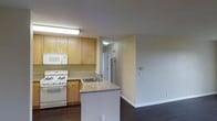 This single-floor space showcases a functional, open layout connecting the kitchen and living area. The kitchen is equipped with light wood cabinetry providing ample storage, a white gas range, built-in microwave, and a stainless steel refrigerator visible in the background. A peninsula with a granite-style countertop extends into the space, offering additional workspace and a natural division between the kitchen and living area. The sink is integrated into the peninsula, adding practicality to the layout. Overhead fluorescent lighting illuminates the kitchen workspace, while a flush ceiling fixture lights the adjacent hallway. Neutral yellow-toned walls create a warm, understated backdrop throughout. Dark hardwood floors run through both the kitchen and adjoining living space, offering a rich contrast to the lighter cabinetry and walls. The open flow between the kitchen and living area enhances the overall sense of space. A hallway visible in the background leads further into the unit, suggesting additional rooms beyond. A wall-mounted thermostat in the living area highlights the practical, well-appointed nature of the space.