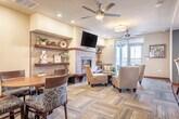 Located at 2400 Parmenter St in Middleton, WI, this single-floor space offers a thoughtfully designed layout that combines functional living with contemporary style. The kitchen is a standout feature, outfitted with rich dark-wood cabinetry extending to the ceiling, stainless steel appliances including a range, dishwasher, and refrigerator, a double stainless sink, and a striking mosaic tile backsplash in cool blue and gray tones. Light wood-style flooring and soft blue accent walls give the kitchen a clean, modern feel, with a window providing natural light and views to an outdoor balcony area. The combined living and dining area is anchored by a tile-clad fireplace with a wall-mounted television above and flanked by open cherry wood shelving. A multi-tone parquet-style carpet adds texture and warmth underfoot, while recessed lighting and ceiling fans illuminate the space efficiently. Glass-panel sliding doors with warm wood frames connect the living area to an adjacent corridor, enhancing openness and flow. A dining nook with a round wood table and upholstered chairs sits alongside accent seating and curated artwork, creating an inviting atmosphere. French doors open to an outdoor balcony, allowing natural light to fill the living area and extending the sense of space beyond the interior.