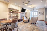 Located at 2400 Parmenter St in Middleton, WI, this single-floor space offers a thoughtfully designed layout that combines functional living with contemporary style. The kitchen is a standout feature, outfitted with rich dark-wood cabinetry extending to the ceiling, stainless steel appliances including a range, dishwasher, and refrigerator, a double stainless sink, and a striking mosaic tile backsplash in cool blue and gray tones. Light wood-style flooring and soft blue accent walls give the kitchen a clean, modern feel, with a window providing natural light and views to an outdoor balcony area. The combined living and dining area is anchored by a tile-clad fireplace with a wall-mounted television above and flanked by open cherry wood shelving. A multi-tone parquet-style carpet adds texture and warmth underfoot, while recessed lighting and ceiling fans illuminate the space efficiently. Glass-panel sliding doors with warm wood frames connect the living area to an adjacent corridor, enhancing openness and flow. A dining nook with a round wood table and upholstered chairs sits alongside accent seating and curated artwork, creating an inviting atmosphere. French doors open to an outdoor balcony, allowing natural light to fill the living area and extending the sense of space beyond the interior.