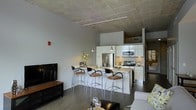 This single-floor space showcases a contemporary urban design highlighted by striking exposed concrete ceilings that lend an industrial character to the interior. The open-concept layout connects the kitchen, dining, and living areas in a seamless, functional flow.

The kitchen is a standout feature, outfitted with crisp white cabinetry, stainless steel appliances including a refrigerator, range, and microwave, and a central island with a light-toned countertop. Pendant lighting hangs above the island, complementing the sleek, modern aesthetic. A set of bar-height stools with chrome frames and warm upholstered seats lines the island, creating a casual dining and gathering space.

Dark hardwood flooring runs throughout, providing a rich contrast to the white cabinetry and light walls. Track lighting along the ceiling adds both functional illumination and a clean, modern design touch. The living area is visible from the kitchen, anchored by a flat-screen television on a wooden media console, and offers a comfortable, open atmosphere. The layout is efficient and well-organized, making excellent use of the available space while maintaining a stylish, cohesive look throughout.