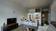 This single-floor space showcases a contemporary urban design highlighted by striking exposed concrete ceilings that lend an industrial character to the interior. The open-concept layout connects the kitchen, dining, and living areas in a seamless, functional flow.

The kitchen is a standout feature, outfitted with crisp white cabinetry, stainless steel appliances including a refrigerator, range, and microwave, and a central island with a light-toned countertop. Pendant lighting hangs above the island, complementing the sleek, modern aesthetic. A set of bar-height stools with chrome frames and warm upholstered seats lines the island, creating a casual dining and gathering space.

Dark hardwood flooring runs throughout, providing a rich contrast to the white cabinetry and light walls. Track lighting along the ceiling adds both functional illumination and a clean, modern design touch. The living area is visible from the kitchen, anchored by a flat-screen television on a wooden media console, and offers a comfortable, open atmosphere. The layout is efficient and well-organized, making excellent use of the available space while maintaining a stylish, cohesive look throughout.