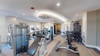 Located at 401 Northeast Mizner Boulevard in Boca Raton, FL, this fitness center offers a thoughtfully designed, single-floor facility with distinct zones catering to a full range of workout needs. The space is organized into several functional areas, beginning with a dedicated cardio section featuring Matrix treadmills, ellipticals, and Peloton cycling bikes arranged along patterned carpet flooring that visually delineates each zone. A strength training area is well-stocked with Matrix cable machines, selectorized equipment, a full dumbbell rack, and benches, providing comprehensive resistance training options. A separate room houses medicine balls, stability balls, and additional functional fitness accessories. The facility is accessed through sleek floor-to-ceiling glass sliding doors with dark metal framing, lending a contemporary, upscale feel. Interior finishes are clean and modern, with neutral walls adorned by framed athletic artwork, recessed lighting, and a distinctive oval tray ceiling detail with ambient lighting in the main training area. Large windows allow natural light to filter in, enhancing the bright and energizing atmosphere. Dual wood-panel entry doors with glass inserts provide additional access. The overall design is polished and professional, creating an inviting environment for residents seeking a premium fitness experience in the heart of Boca Raton.