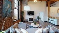 This single-floor urban loft is a masterful fusion of historic industrial architecture and contemporary living. The living area commands attention with its soaring ceilings adorned with original exposed wooden beams and visible ductwork, reinforcing the space's industrial character. Oversized arched windows line the exposed brick walls, flooding the room with natural light and offering sweeping panoramic city views. A sleek fireplace anchors the living area, complemented by hardwood flooring throughout.

The kitchen is well-appointed with light maple cabinetry, granite countertops, black appliances including a range, built-in microwave, dishwasher, and refrigerator, along with ample counter and storage space. A galley-style layout maximizes functionality, with a peninsula providing additional prep space.

The generously sized bedroom continues the loft's signature aesthetic, with exposed brick walls, tall multi-pane windows framing elevated city views, and soaring ceilings with wooden beam detailing. A ceiling fan adds comfort to the expansive room, which includes closet space and room for a seating area.

The bathroom features a dual-sink vanity with a dark granite countertop, light maple cabinetry, a large wall mirror, and bright overhead lighting, offering a clean and practical design. Throughout, the space balances raw architectural character with modern finishes.