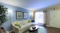 This single-floor space offers a warm and inviting atmosphere, highlighted by a thoughtfully designed living room. The room is defined by soft blue accent walls that create a calm, relaxing ambiance, complemented by crisp white crown molding that adds a touch of classic elegance. A large window dressed with neutral linen-style curtains and horizontal blinds allows abundant natural light to fill the space, creating an airy and open feel. The hardwood flooring anchors the room with a rich, warm tone that contrasts beautifully with the cool wall color. The living area comfortably accommodates a spacious sectional sofa in a light cream tone, accented with green and blue decorative pillows that echo the room's color palette. A dark wood oval coffee table serves as a stylish centerpiece, while a coordinating area rug adds texture and definition to the seating area. Two framed nature-inspired artworks adorn the blue wall, contributing to the serene, nature-influenced aesthetic of the space. A floor lamp provides additional ambient lighting, enhancing the room's cozy character. Overall, the space is cohesive, well-lit, and designed to provide comfort and style in equal measure.