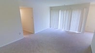 This single-floor space offers a clean, functional layout designed for comfortable everyday living. The main living area is generously sized, featuring soft, neutral-toned wall-to-wall carpeting that adds warmth underfoot. A large set of floor-to-ceiling windows dressed with vertical blinds lines one wall, allowing abundant natural light to fill the room and creating a bright, airy atmosphere throughout the day. The crisp white walls enhance the sense of openness, providing a versatile backdrop for a variety of furnishing styles. A clearly defined entryway or adjoining hallway connects to the rest of the space, offering a natural flow between areas. Recessed ventilation and clean architectural lines contribute to the space's understated, well-maintained character. The overall ambiance is light, neutral, and move-in ready, making it an ideal canvas for personalization. The combination of ample natural light, open floor space, and practical layout gives this home a welcoming and comfortable feel throughout.