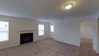 This single-floor space presents a clean, move-in-ready living area finished in a neutral, light color palette throughout. The main living room is spacious and carpeted in warm beige tones, providing a comfortable and inviting foundation. A black-framed fireplace with a white mantel serves as the room's focal point, adding both a functional heating element and a classic architectural detail. Two windows flank a paneled entry door, allowing natural light to flow into the space and creating a bright, airy atmosphere. The flush-mounted ceiling light fixture ensures consistent illumination throughout the room. Smooth white ceilings and crisp white trim complement the soft wall tones, contributing to a cohesive and understated aesthetic. The overall layout is open and straightforward, offering flexibility for a variety of furniture arrangements. The entry door features a glass panel insert, allowing additional light to filter in while maintaining a traditional residential look. The space reflects a well-maintained, practical design suited for comfortable everyday living.