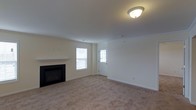 This single-floor space presents a clean, move-in-ready living area finished in a neutral, light color palette throughout. The main living room is spacious and carpeted in warm beige tones, providing a comfortable and inviting foundation. A black-framed fireplace with a white mantel serves as the room's focal point, adding both a functional heating element and a classic architectural detail. Two windows flank a paneled entry door, allowing natural light to flow into the space and creating a bright, airy atmosphere. The flush-mounted ceiling light fixture ensures consistent illumination throughout the room. Smooth white ceilings and crisp white trim complement the soft wall tones, contributing to a cohesive and understated aesthetic. The overall layout is open and straightforward, offering flexibility for a variety of furniture arrangements. The entry door features a glass panel insert, allowing additional light to filter in while maintaining a traditional residential look. The space reflects a well-maintained, practical design suited for comfortable everyday living.