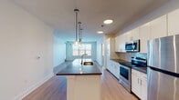 This well-appointed two-bedroom residence offers a thoughtfully designed single-floor layout that balances modern aesthetics with everyday functionality. The open-concept living and kitchen area serves as the heart of the home, anchored by a central island with a dark countertop and pendant lighting overhead. The kitchen is outfitted with crisp white cabinetry, grey subway tile backsplash, dark countertops, and a full suite of stainless steel appliances including a range, microwave, and refrigerator. Light wood-style flooring flows throughout the main living areas, enhancing the bright, airy atmosphere created by large windows that invite generous natural light. The spacious living area is served by a ceiling fan and offers an open sightline to the kitchen. Both bedrooms feature plush carpeting, ceiling fans with integrated lighting, and windows framing urban views. One bedroom includes direct access to a private balcony. The unit is complemented by two bathrooms—a full bath featuring a white vanity with dark countertop, tub/shower combo, and light wood-style flooring, as well as a second bathroom with a single-sink vanity and modern fixtures throughout. Clean white walls and a neutral palette create a cohesive, contemporary feel across the entire residence.